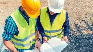 Two construction workers wearing safety helmets and high-visibility vests reviewing building plans on site.