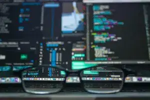 Computer screen with colorful code and system interfaces viewed through eyeglasses on a keyboard.