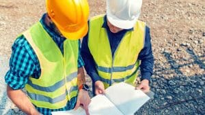 Two construction workers wearing safety helmets and high-visibility vests reviewing building plans on site.