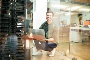 Smiling IT professional with laptop working in a modern office server room.